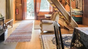 A rustic cabin interior with a wooden staircase, a sofa, a table, and a view of the outdoors through large windows.