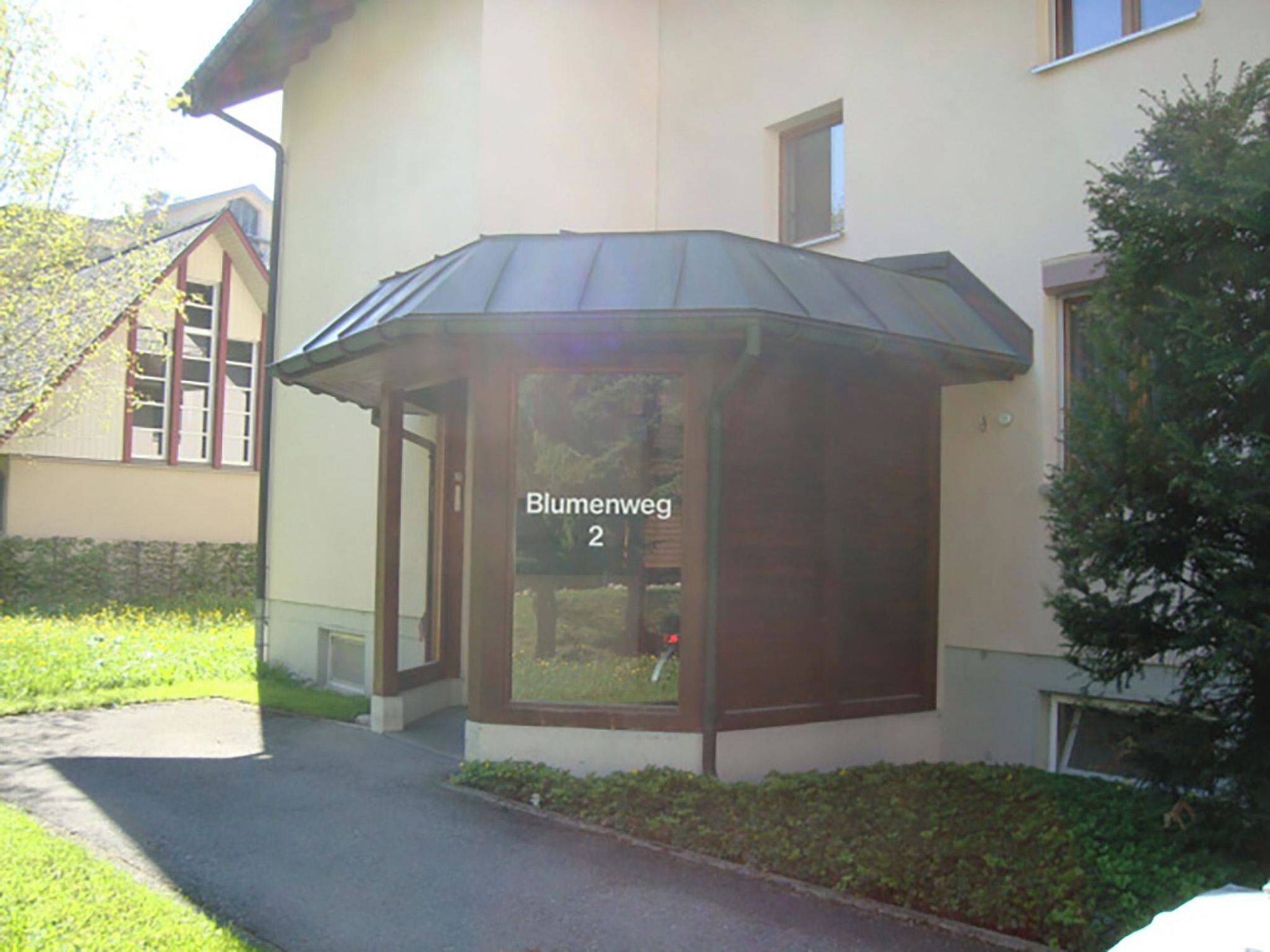 A building with a corner entrance, a sign reading 'Blumenweg 2', and a small garden area.