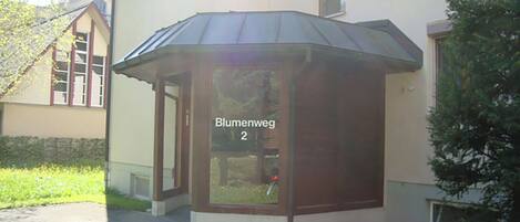 A building with a corner entrance, a sign reading 'Blumenweg 2', and a small garden area.