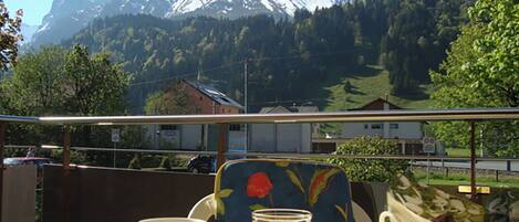 A table with a cup, a glass of orange juice, and a basket of bread, overlooking a snow-capped mountain and a forested valley.