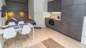 A compact living space with a bed, a dining table with chairs, a kitchenette with dark cabinets, and a white door.