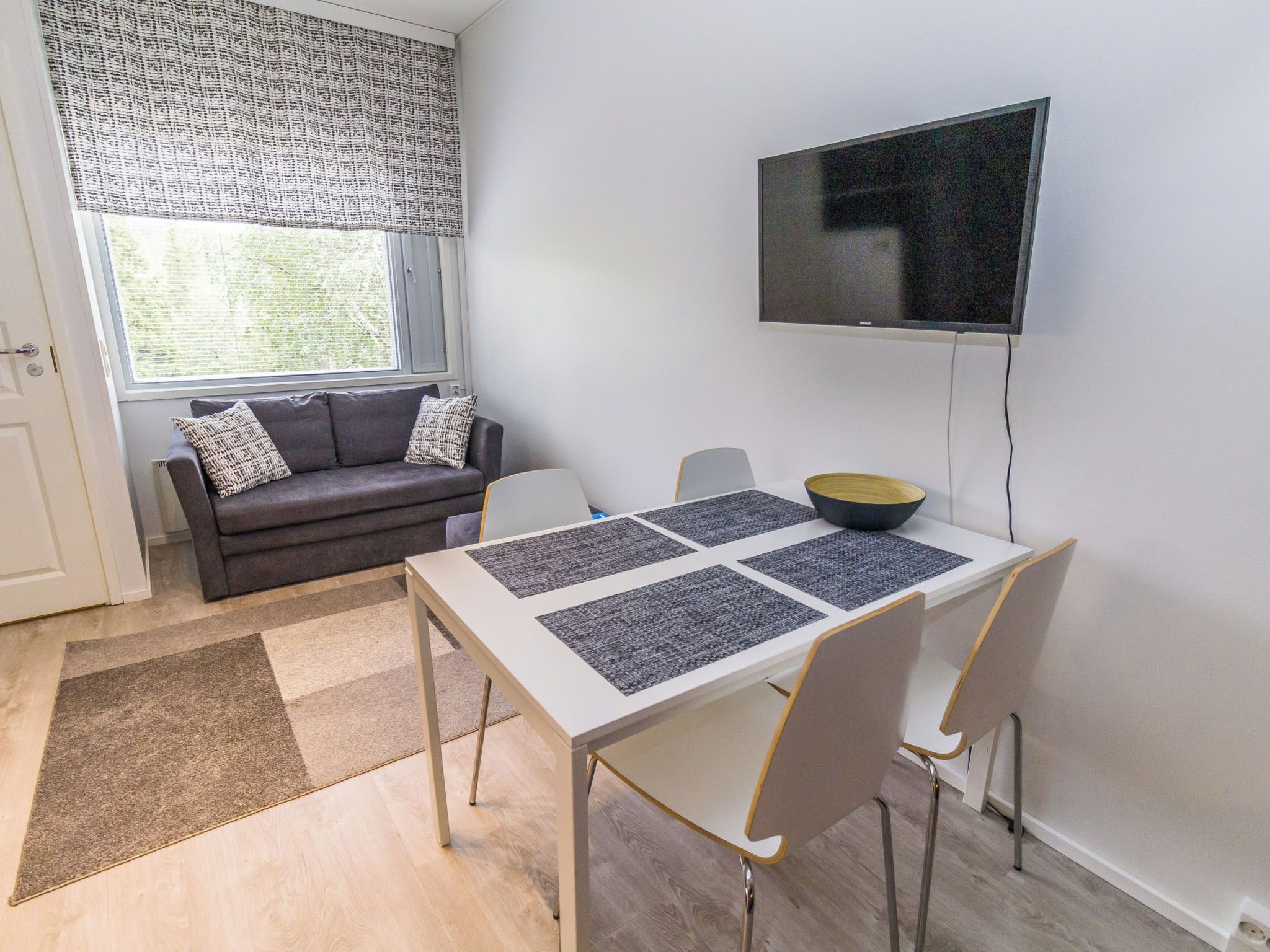 A modern living room with a sofa, a dining table with placemats, a television, and a window with blinds.