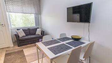 A modern living room with a sofa, a dining table with placemats, a television, and a window with blinds.