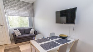 A small, modern room with a sofa, a dining table with four chairs, a TV mounted on the wall, and a window with blinds.