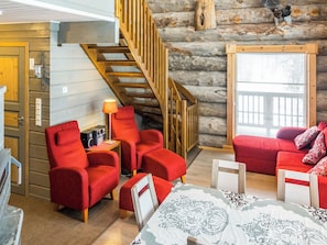 A log cabin interior with a staircase, red furniture, a table with a lace tablecloth, and a window with blinds.
