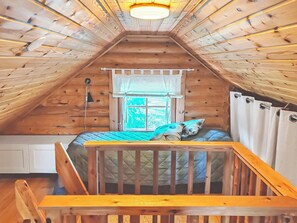A loft bedroom with a wooden ceiling, a window with a view, a bed with a quilt, and a small table.