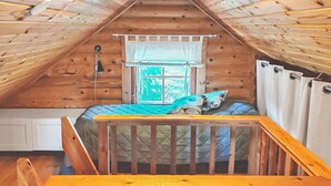 Ein gemütliches Schlafzimmer im Dachgeschoss mit schrägverlegtem Holzboden, einem Bett mit gesteppter Decke, einem Fenster mit Vorhängen und einem kleinen Tisch.