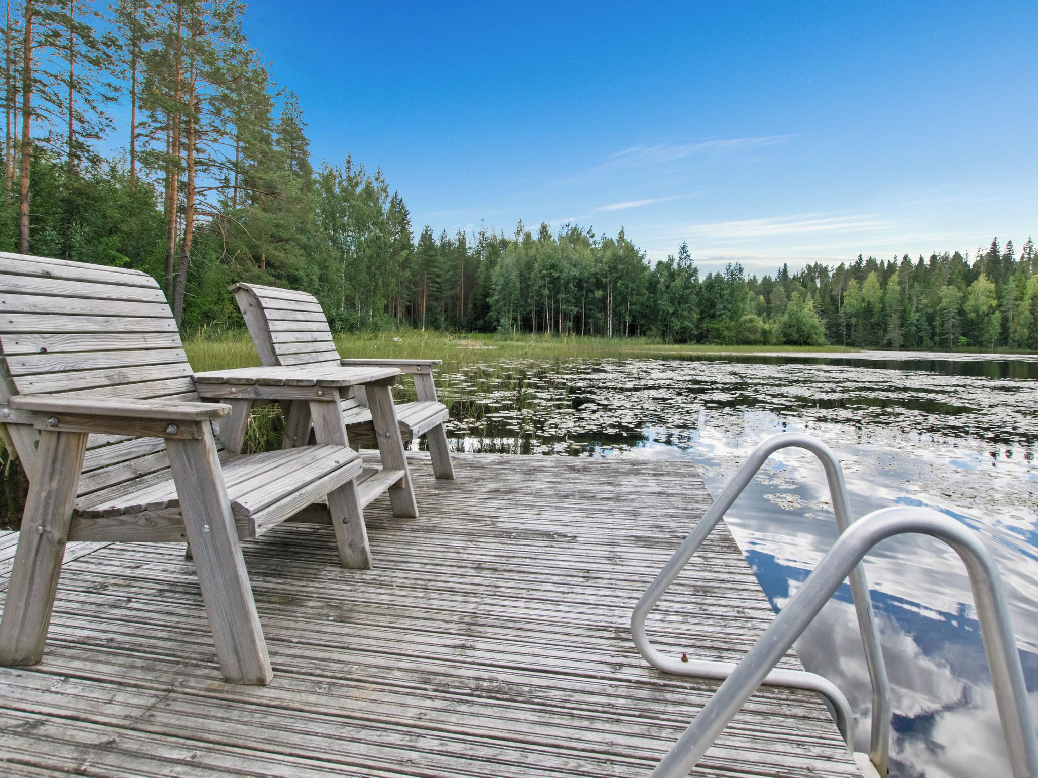 A wooden deck with two chairs and a table, overlooking a lake with lily pads and surrounded by trees.