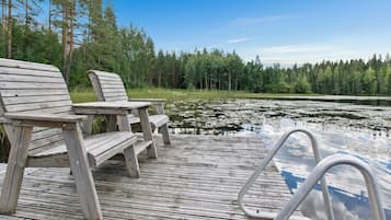 A wooden deck with two chairs and a table, overlooking a lake with lily pads and surrounded by trees.