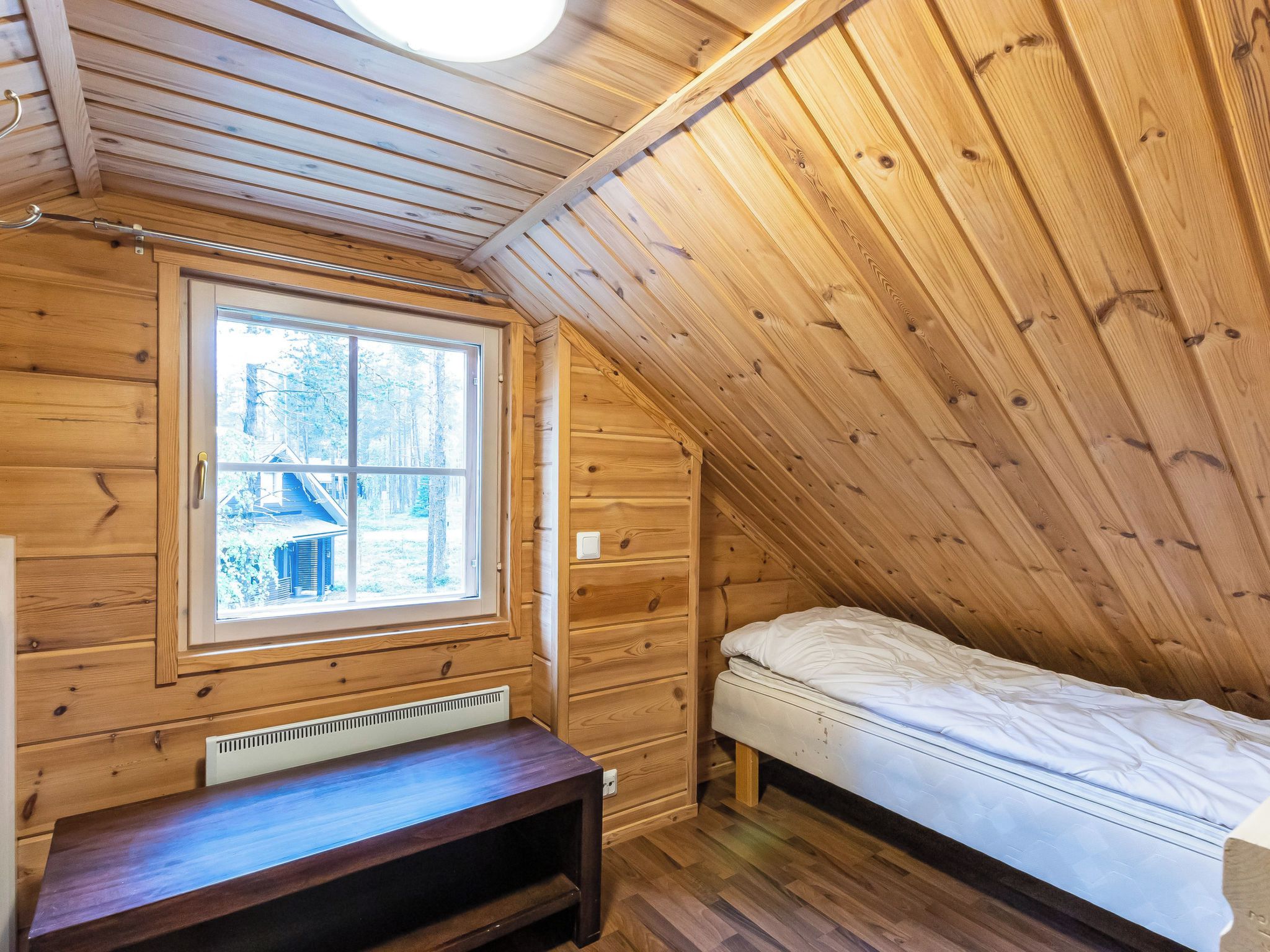 Una habitación pequeña de madera con techo inclinado, una cama individual, un banco y una ventana con vista a una casa y árboles.