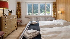 Camera d'albergo con un letto, una cassettiera in legno, un termosifone e una finestra vista foresta e edifici.
