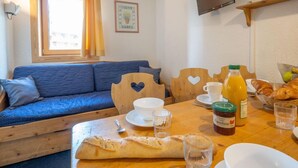 Een knus vertrek met een blauwe bank, houten stoelen, een eettafel met ontbijtartikelen en een aan de muur bevestigde televisie.