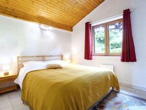 A bedroom with a wooden ceiling, a bed with a yellow blanket, a nightstand with a lamp, and a window with red curtains.