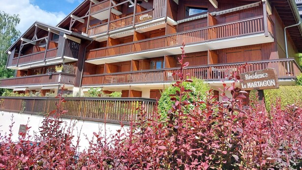 Un edificio de madera de varios pisos con balcones, un cartel que dice "Residence Le Martagon" y un jardín bien cuidado con plantas de hojas rojas.