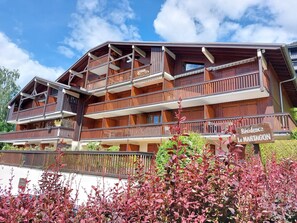 A multi-story wooden building with balconies, a sign reading "Residence Le Martagon," and a well-maintained garden with red-leafed plants.