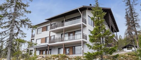 A modern multi-story building with balconies, surrounded by trees and rocky terrain.