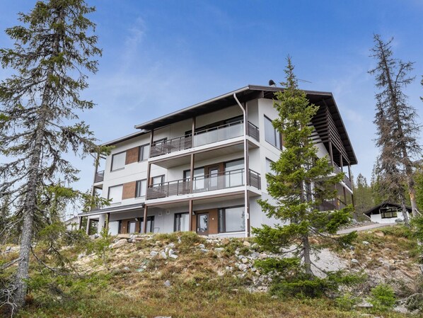 A modern multi-story building with balconies, surrounded by trees and rocky terrain.