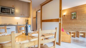 A wooden interior with a dining table, chairs, a kitchen area with a microwave, and a wooden wall with a picture. - Arcelle 203 by Interhome (Val Thorens)