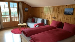 A room with wooden walls, a bed with red bedding, a grey sofa with cushions, a small round table, and a window with a view of mountains. - Apartment Hari by Interhome (Adelboden)