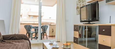 A hotel room with a bed, a TV, a wooden cabinet, a table with cups and pastries, and a balcony with chairs.