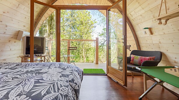 A cozy room with a bed, a TV, a table with a chessboard, and a view of trees through an open archway. - Monchateau étoilé Hébergement insolite (Merlas)