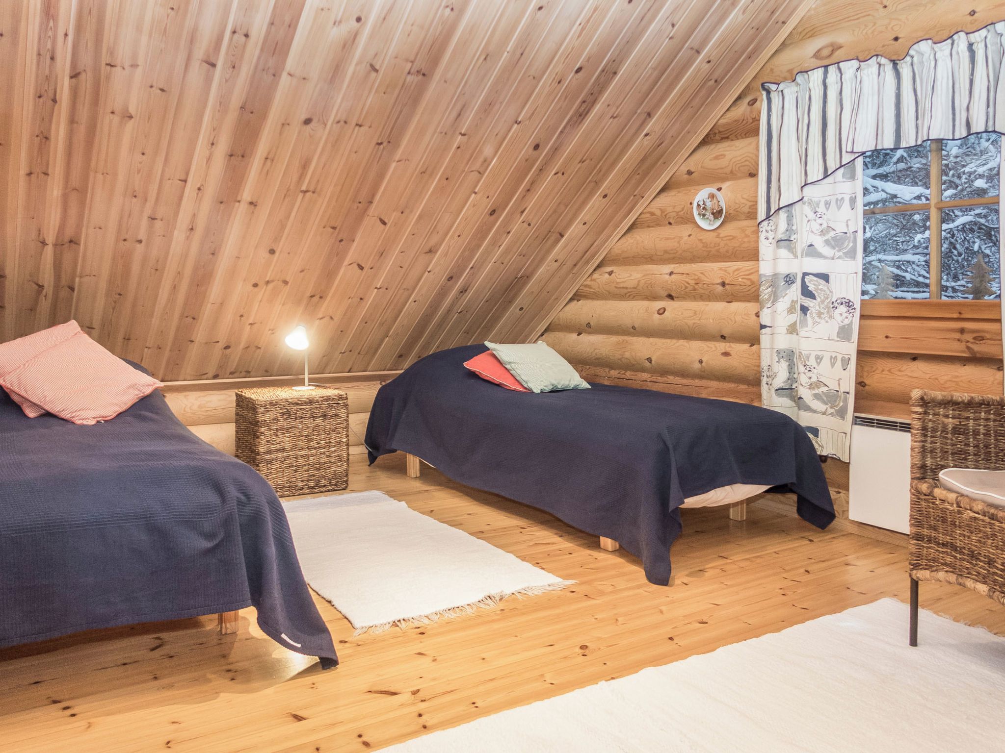 A cozy cabin with two beds, a small table, a lamp, and a window with a view of a snowy landscape.