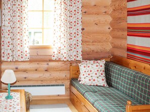 Interior d'una cabana acollidora amb una paret de fusta, una finestra amb cortines florals, un llit amb roba de llit a quadres i una làmpada sobre una taula.
