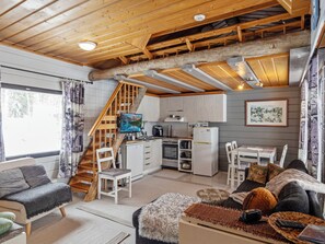 Interior acogedor de una cabaña con escalera de madera, zona de cocina, mesa de comedor y sala de estar con sofá y chimenea.