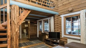 Interior de una cabaña de troncos con escalera de madera, televisor, sofá y vistas al exterior.