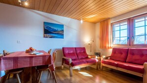 Una sala de estar acogedora con techo de madera, una mesa de comedor con mantel rojo, un sofá rojo y un ventanal con cortinas.