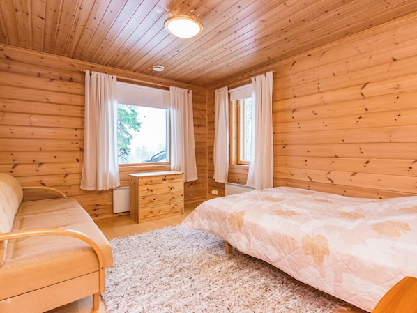 A wooden cabin bedroom with a bed, a chair, a dresser, and two windows with curtains.