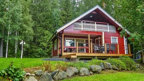Una cabaña roja con terraza de madera y porche cubierto, rodeada de un paisaje verde exuberante y árboles.