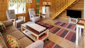 Interior de una cabaña de troncos con un sofá, sillas, un televisor, una escalera y un ventanal con cortinas.