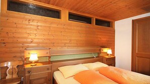 Una habitación de cabaña de madera con una cama, dos mesitas de noche, una silla y una ventana.