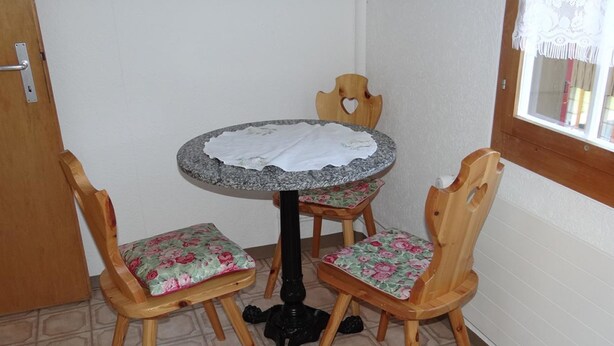 Een kleine ronde tafel met een wit kleed, twee houten stoelen met bloemenkussens, en een zwart onderstel.