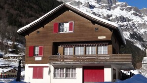 Een chalet met een houten gevel, rode luiken en een balkon met uitzicht op een besneeerd berglandschap.