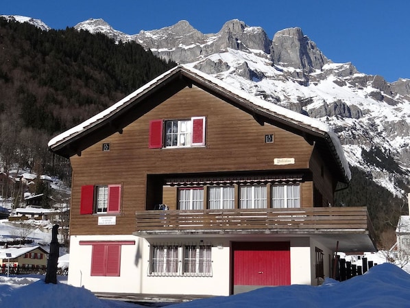 A chalet with a wooden facade, red shutters, and a balcony overlooking a snowy mountain landscape.