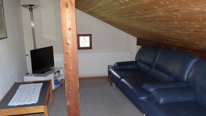A living room with a blue sofa, a television on a stand, a wooden table with a lace tablecloth, and a wooden ceiling beam.