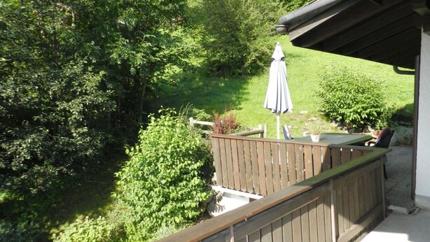 Une terrasse en bois avec une table et des chaises, entourée d’une végétation luxuriante et agrémentée d’un parasol.