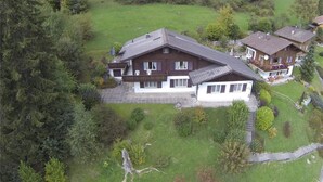 Une grande maison avec un toit en pente, entourée d’une pelouse bien entretenue et d’une forêt dense.