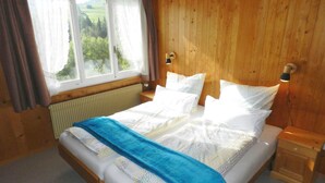 Une chambre à coucher aux murs lambrissés, avec un lit, deux tables de chevet, une fenêtre avec des rideaux et une vue sur la verdure à l’extérieur.