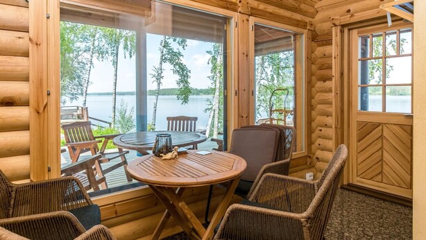 Une cabane en bois avec une table ronde et des chaises en osier, de grandes fenêtres donnant sur un lac et une forêt.