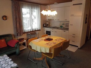 Eine kompakte Küche mit weißen Schränken, einem Esstisch mit Blumenmuster-Tischdecke und einem Sofa mit rotem Kissen.