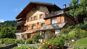 Eine zweistöckige Holzhütte mit Balkon, umgeben von einem Garten mit verschiedenen Blumen.