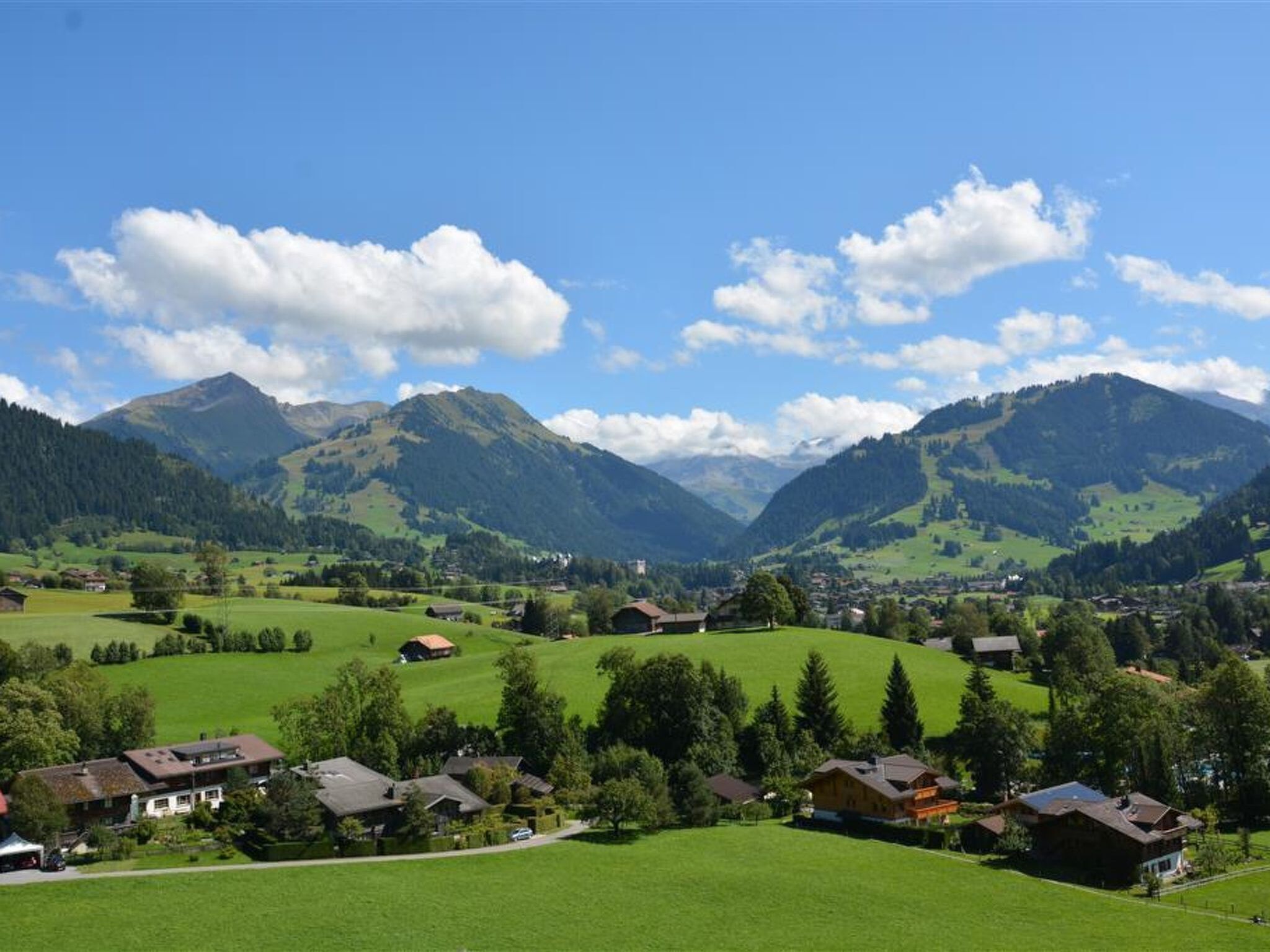 Un paysage rural avec des maisons, des champs verdoyants et des montagnes sous un ciel dégagé.