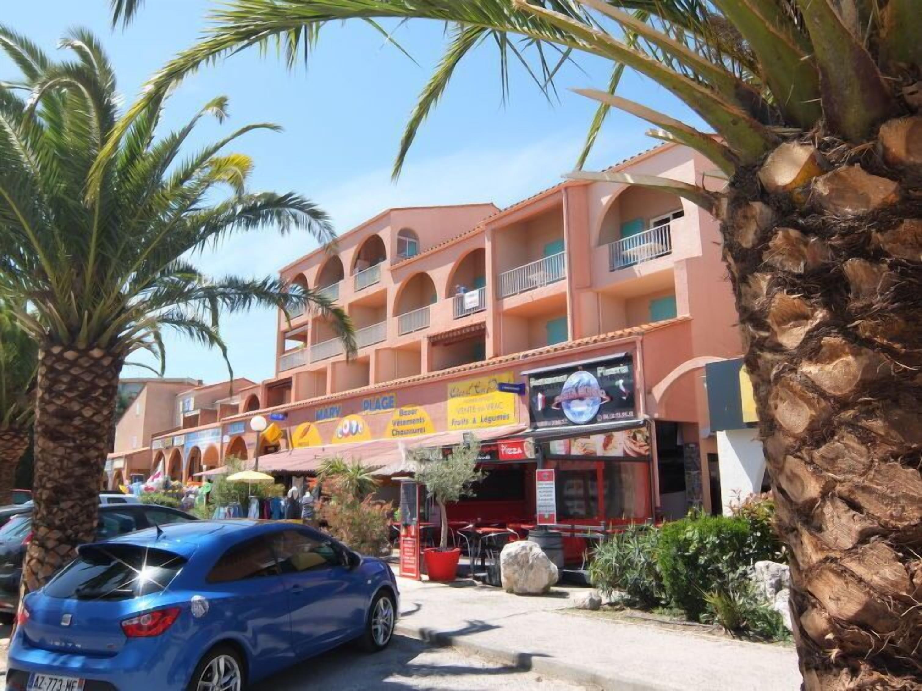 A multi-story building with balconies, palm trees, and a blue car parked in front.