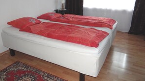Una cama doble con ropa de cama roja, una mesita de noche pequeña con una lámpara, un cuadro en la pared y una cortina oscura.