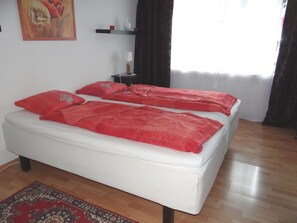 Un letto matrimoniale con biancheria rossa, un comodino con una lampada, un quadro al muro e una tenda scura.