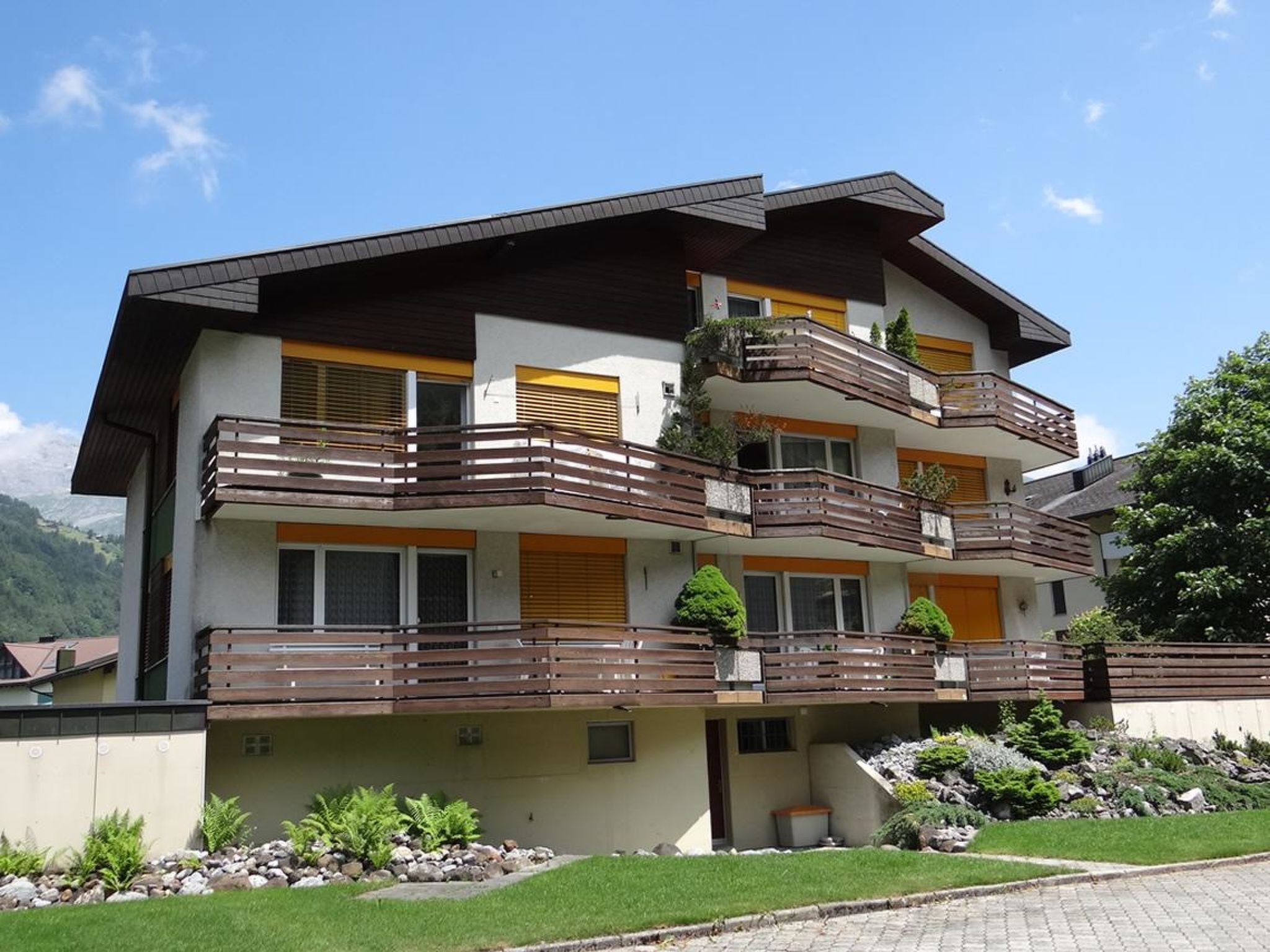 A multi-story residential building with balconies, a stone facade, and a mountainous backdrop.