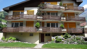 Un edificio de varios pisos con balcones, una puerta roja y un paisaje montañoso de fondo.